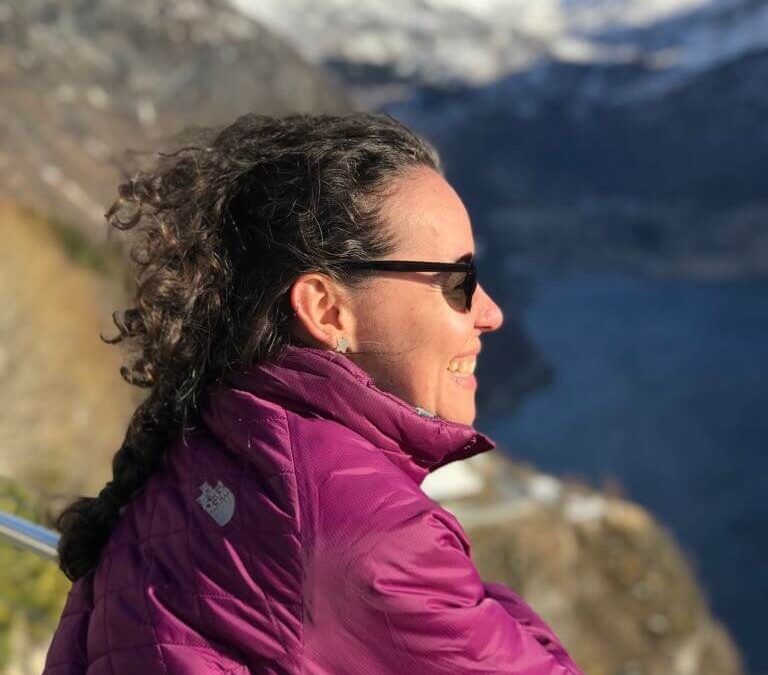 Mulher de posa de lado, apoiada em uma grade baixa, sorrindo e olhando para o horizonte a sua frente. Ao fundo, os fiordes noruegueses, formados entre montanhas.