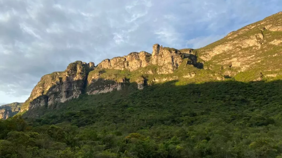 Montanha se ergue no Vale do Pati. O sol reflete sobre a formação rochosa, enquanto o céu está um pouco nublado e abaixo há vegetação verde.