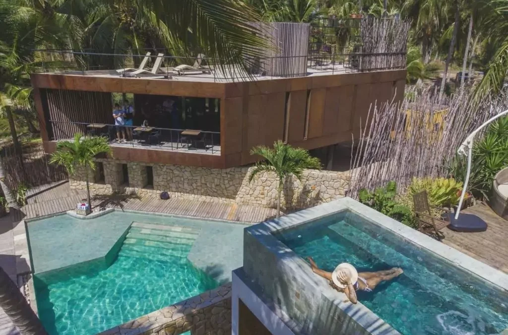 Hotel Pedras do Patacho, na Praia do Patacho, Alagoas. Uma construção em meio à vegetação, com uma piscina no centro, em que uma mulher de chapéu toma sol.